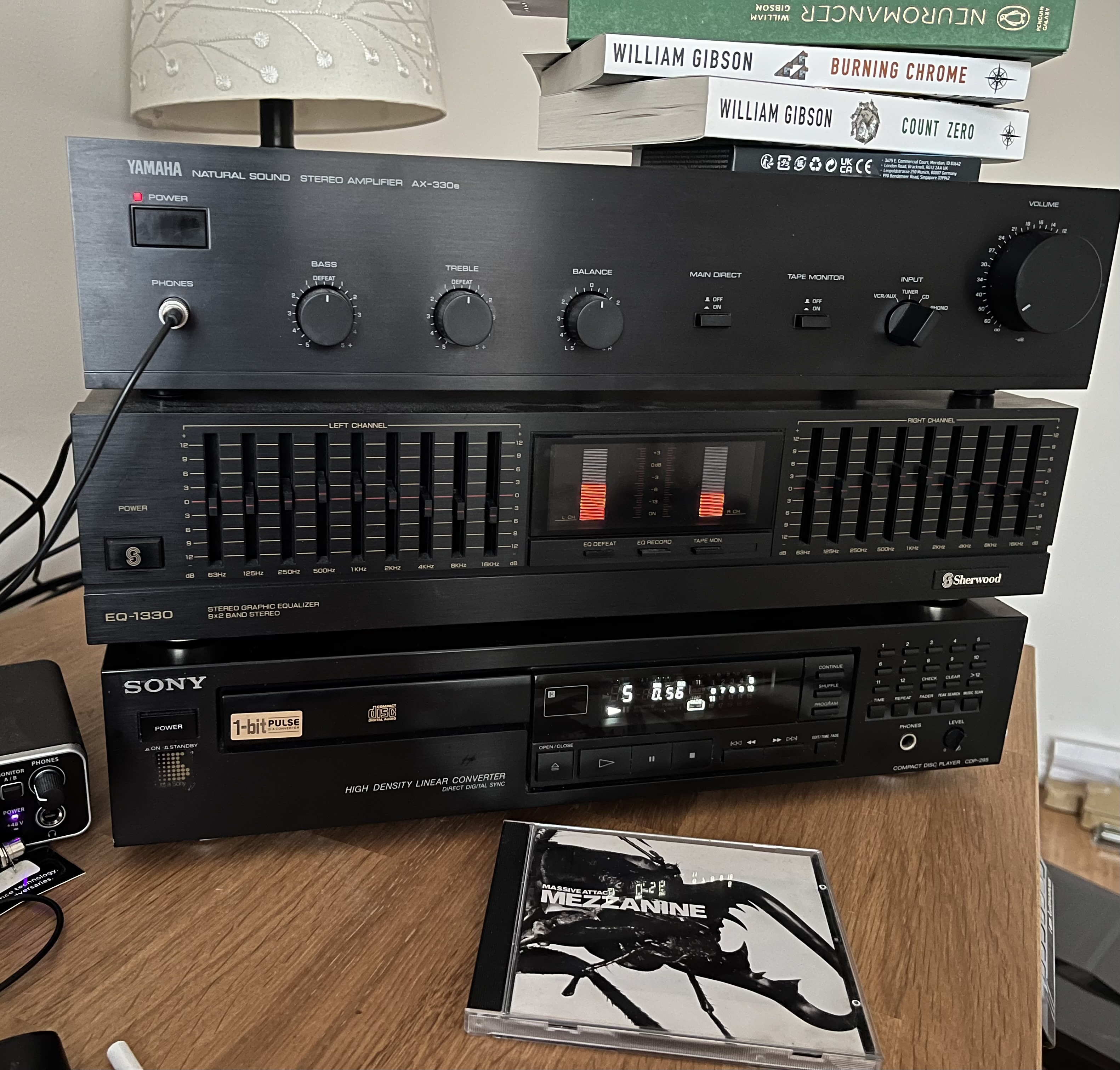 A stack of HiFi equipment, on a desk. The top of the stack has three books on top - Neuromancer, Burning Chrome and Count Zero by William Gibson. The top unit is a Yamaha AX-330e, which is powered on. It has a volume knob, input selection dial, main direct and tape monitor switch, and 3 smaller dials labelled Bass, Treble and Balance. Headphones are plugged in. Below it, is the EQ-1330 previously mentioned in this post, and it's powered on and shows two VU meters illuminated. Below this, is the Sony CD-295, playing track 5 of Mezzanine by Massive Attack. The CD case for Mezzanine is in front of the CD player.